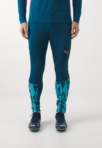 Männer Sportleggings in Teal mit einem geometrischen blauen Muster an den Unterschenkeln, versehen mit einem pinkfarbenen Logo auf dem Oberschenkel. Geeignet für Athletikbekleidung.