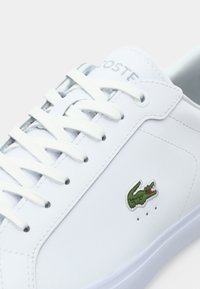 Baskets en cuir blanc avec des lacets blancs et un logo crocodile vert sur le côté, montrant une partie de la languette avec le nom de la marque.
