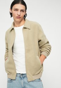 Giacca in pile beige con chiusura a zip e finitura testurizzata, dotata di colletto, polsini a costine e tasche laterali, indossata su una t-shirt bianca.