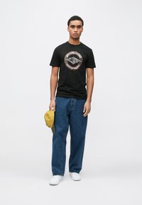 Sort t-shirt med et grafisk logo, blå jeans og hvide sneakers. Personen holder en gul kasket med en grå skygge. Simpelt baggrund.