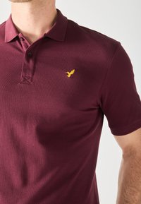 Burgunderrød polo skjorte lavet af tekstureret stof med en to-knaps placket og en lille gul fuglebroderi på brystet.