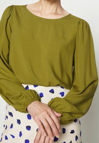 Blouse vert olive avec des manches bouffantes et des poignets élastiques, associée à une jupe crème avec un motif de taches bleues. Texture de tissu lisse.