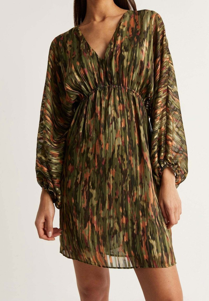 Femme portant une robe mini plissée à manches longues avec un motif abstrait vert, orange et marron et une taille cintrée.