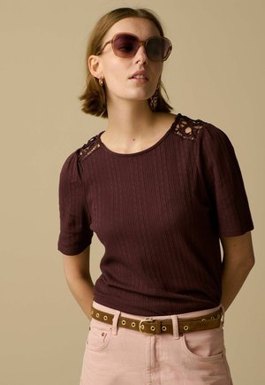 Woman wearing large brown sunglasses, a maroon short-sleeve top with lace details on shoulders, light pink pants, and a brown belt.