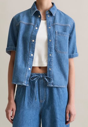 Osoba w niebieskiej dżinsowej koszuli z podwiniętymi rękawami, założonej na białą, krótką prążkowaną koszulkę na ramiączkach, w połączeniu z wysokim stanem dżinsowych spodni przewiązanych kokardą.