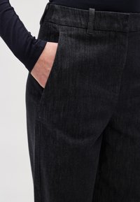 Tumša džinsa bikses ar gludu tekstūru, kurām ir sānu kabatas un pieskaņots piegriezums. Vidukļa josta ir aprīkota ar jostas cilpām.