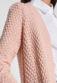 Mujer vistiendo un cárdigan de punto texturizado rosa claro sobre una camisa blanca con cuello, mostrada de hombro a cintura sobre un fondo blanco.
