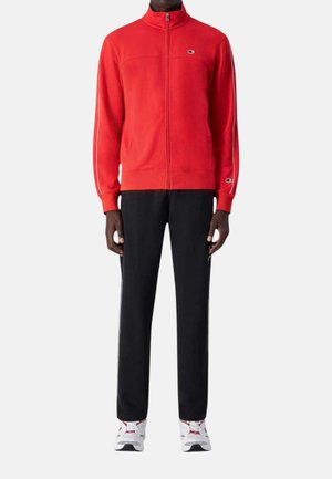 Uomo che indossa una giacca rossa con zip, pantaloni neri e sneakers bianche, in piedi davanti a uno sfondo chiaro e uniforme.
