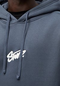 Mikina s kapucí v tmavě modré barvě, se bílým vyšitým logem na hrudi a šňůrkami s texturovanými konci.