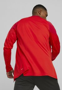 Rotes Sportshirt mit langen Ärmeln, strukturiertem, einfarbigem Körper und gemusterten Ärmeln. Mit abgerundetem Saum und einem seitlichen Reißverschlussdetail nahe dem unteren Ende.