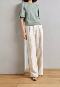 Grøn bomulds sweatshirt med pufærmer, parret med beige linnedragt. Polstret skulderdetalje og vid ben-design. Lys træbund.