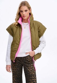 Olivově zelená prošívaná vesta s růžovou podšívkou a zipem, s krátkými rukávy a nastavitelným lemem. Nošena přes bílou košili a kalhoty s leopardím potiskem.