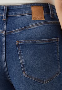 Tunna blå denimjeans med en läderetikett märkt "PIECES." Har bakfickor och orange sömnad för att ge extra detaljer.