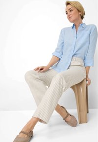 Helles blaues Hemd mit Knopfleiste, beige gemusterte Hose und taupefarbene Ballerinas. Der Stuhl ist aus glattem, gebogenem Kunststoff in einem hellbraunen Farbton.