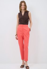 Brown sleeveless top with buttons, paired with coral tailored pants. Features a smooth texture and a slim fit. Worn with brown heeled sandals.