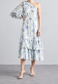 Robe florale à une épaule en blanc et bleu, avec des accents froncés, un ourlet à niveaux, et une taille cintrée avec un détail à nouer. Texture de tissu douce.