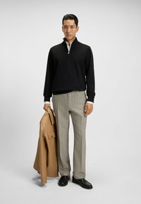 Maglione nero con zip, pantaloni a quadri, tiene in mano un giubbotto di pelle beige e indossa scarpe nere. Tessuto liscio e vestibilità su misura.