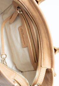Binnenkant van een tan leren handtas met een ritszakje en een merklabel. Glad oppervlak met een lichtbeige voering en zichtbare stiksels.