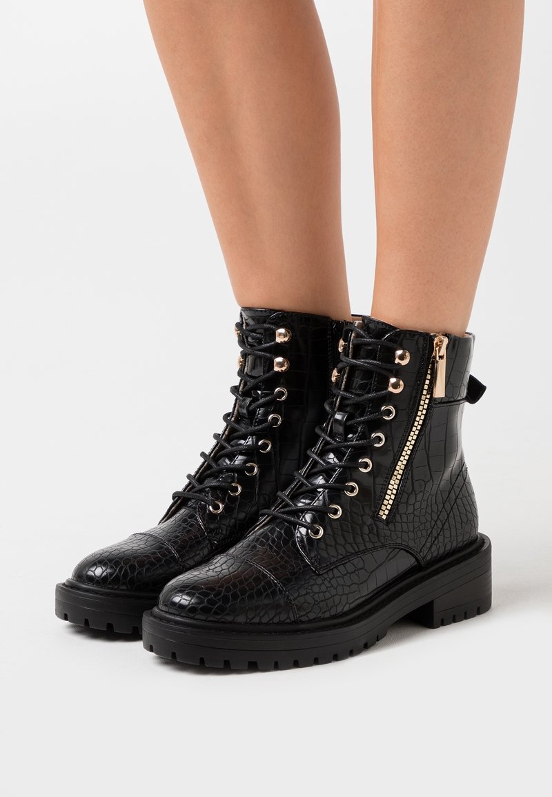 Bottes noires en cuir à texture crocodile, lacées, avec fermeture éclair latérale dorée et semelles épaisses en caoutchouc, portées sur des jambes nues.