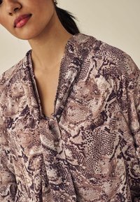 Blouse, millel beež ja pruun sissetungimismustriga, pehme ja tekstuurse kangaga; lahtine lõige ja seotud kaeluse detail.