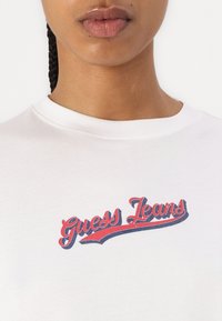 Valkoinen puuvillapaita, jossa on "Guess Jeans" -logo punaisena ja sinisenä graafisena printtinä. Pyöreä pääntie ja lyhyet hihat, sileä pinta.