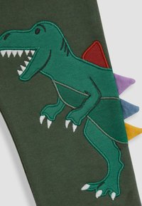 Zielony materiał z haftem przedstawiającym dinozaura T-rexa, z białymi zębami oraz kolorowymi kolcami w odcieniach fioletu, czerwieni i żółci wzdłuż krawędzi.