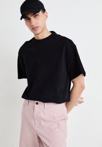 T-shirt noir à manches courtes en coton, associé à un pantalon rose clair avec une fermeture à bouton à l'avant et un design minimaliste. Casquette noire incluse.