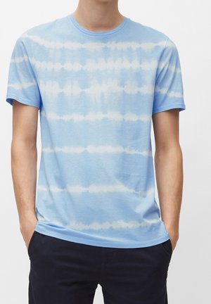 Man die een lichtblauw T-shirt met korte mouwen draagt, voorzien van witte horizontale tie-dye strepen, en donkere broek, met handen in de zakken, staand tegen een effen achtergrond.