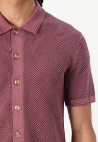 Korte mouw polo in diep mauve, met een gestructureerd breipatroon, klassieke kraag en houten knopen voor de sluiting.