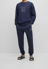 Sudadera navy con el logotipo "HUGO", pantalones navy a juego y sandalias verdes con el texto "HUGO". Estilo casual, ajustado relajado con puños elásticos.
