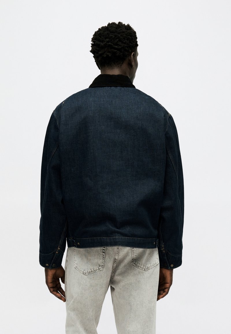 Hombre con cabello corto y rizado, vistiendo chaqueta de mezclilla azul oscuro y jeans gris claro, de espaldas sobre un fondo blanco liso.