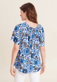 Kurzärmeliges Bluse in blauem und schwarzem Blumenmuster, mit gerafftem Ausschnitt und weichem Stoff. Kombiniert mit weißen Hosen, die einen lockeren Schnitt haben.
