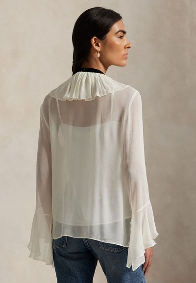 Balta, viegla blūze ar plīsētu apkakli un uzliesmotām piedurknēm, ar caurspīdīgu tekstūru un smalkām vertikālām līnijām, kombinēta ar džinsa biksēm.