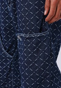 Pantaloni in denim blu con un motivo geometrico di cucitura bianca, dotati di tasca laterale e cuciture a contrasto. Il tessuto ha un aspetto testurizzato.