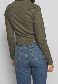 Oliivivihreä denim-takki, jossa on istuva muotoilu, nepparikoristeet ja hennot tikkausdetailit, yhdistettynä korkeavyötäröisiin sinisiin farkkuihin.