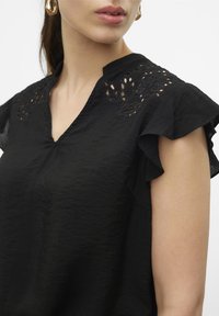 Blusa nera con scollatura a V, caratterizzata da maniche voluminose e delicati ricami floreali sulle spalle. Realizzata in un tessuto leggero e texturizzato.