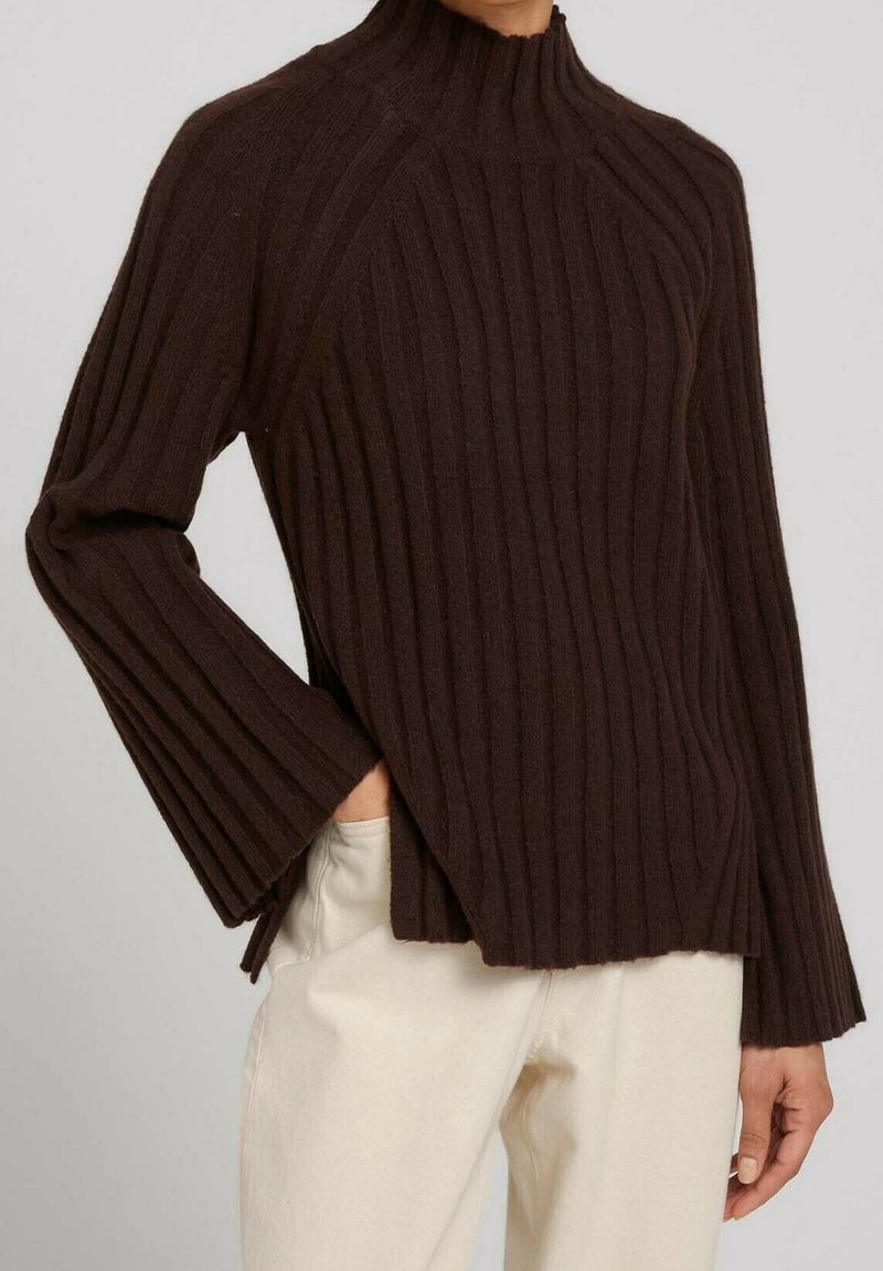 Ciemnobrązowy sweter prążkowany z wysokim dekoltem, długimi poszerzanymi rękawami i bocznymi rozcięciami, noszony z jasnobeżowymi spodniami.