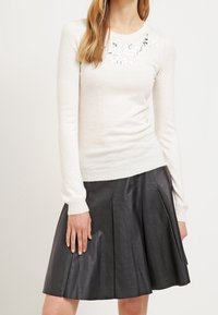 Pull ajusté beige clair avec broderie florale au niveau du col, associé à une jupe plissée en cuir noir, mettant en valeur des textures lisses.