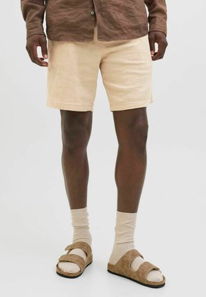 Beige textuur shorts met een middellange snit tot boven de dij, gecombineerd met tan suedine sandalen en crèmekleurige sokken. Het model draagt een bruin shirt met lange mouwen.