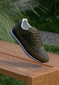 Olivgrüne Sportschuhe mit strukturierten Einsätzen, einem weißen gepolsterten Kragen und einer schwarzen Sohle, auf einer Holzoberfläche präsentiert.