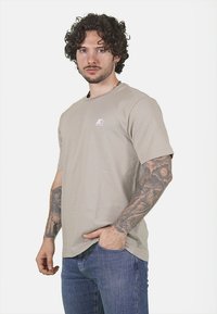 T-shirt grigio chiaro a maniche corte in cotone, con un piccolo logo bianco sul lato sinistro del petto, abbinato a jeans blu.