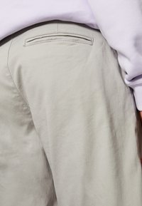 Un pantalon gris clair présente un tissu lisse et texturé avec une poche arrière et un détail de couture subtil. Un sweat-shirt lavande est partiellement visible au-dessus.