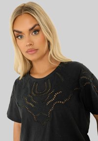 Mustassa lyhythihaisessa t-paidassa on monimutkaisia leikattuja muotoiluja ja brodeerauksia kaula-aukolla; pehmeä rakenne ja rento istuvuus.