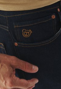 Jeans in denim blu scuro con un logo dell'orsa dorata ricamato sulla tasca anteriore, con cuciture gialle a contrasto e una texture liscia.