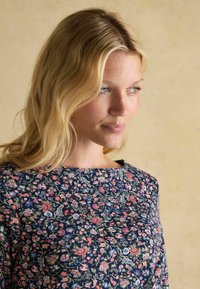 Modrá tkanina s kvetinovým vzorom v ružovej a krémovej farbe, s širokým výstrihom a textúrovaným povrchom, nosená osobou s vlnitými vlasmi.