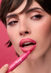 Marcador de labios de doble punta rosa sostenido cerca de los labios, con un acabado brillante, una punta aplicadora redondeada y la marca audaz "Vamp!" en el cuerpo.