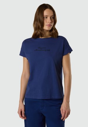 Donna con capelli castani di media lunghezza indossa una maglietta a maniche corte blu navy e pantaloni abbinati, guarda verso destra con un'espressione neutra.