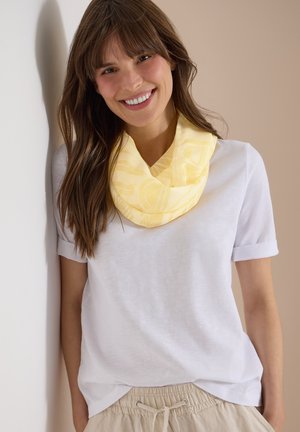 Femme aux longs cheveux bruns souriante, portant une écharpe jaune à motifs, un haut blanc à manches courtes et un pantalon beige à cordon, appuyée contre un mur.