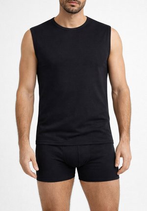 Uomo con maglietta nera senza maniche e boxer neri in piedi contro uno sfondo bianco.