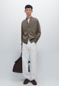 Brun stickad cardigan med hög hals, svarta knappar, i kombination med vita byxor och bruna snörskor. Modellen håller en mörkbrun väska.